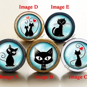 Pode incluir: Cinco botões de armário diferentes com fundo azul e branco e silhuetas de gatos pretos. Os botões estão em diferentes acabamentos: ouro, prata, preto e cinza. Os gatos estão em diferentes poses, incluindo um gato com um coração, um gato com o rabo enrolado e um gato com um coração acima da cabeça. Os botões estão etiquetados com as letras A a E.