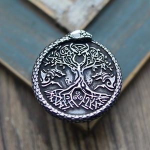 May include: A silver pendant featuring a detailed tree of life design with a snake wrapped around the trunk. The pendant is circular and has a textured surface.