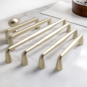 May include: A collection of brushed gold cabinet hardware, including handles and a knob. The handles have a sleek, modern design with a curved shape. The hardware is arranged on a white marble surface, showcasing the metallic finish and design.