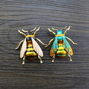 Op de afbeelding: Twee gouden bijen broche met zwarte en gele strepen. E&eacute;n bij heeft witte vleugels en de andere blauwe vleugels.