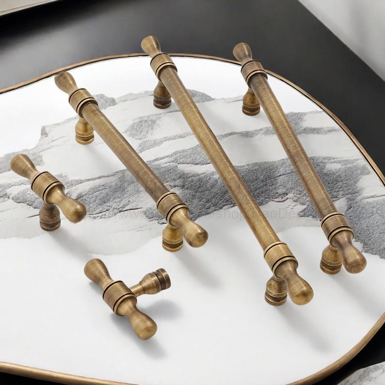 May include: A selection of antique brass cabinet handles and knobs. The image shows long bar pulls and smaller T-shaped knobs, displayed on a white and grey marble-effect surface. The brass hardware has an aged finish, highlighting the detailed design.