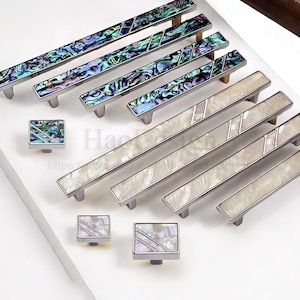 Pode incluir: Uma coleção de ferragens para armários, incluindo puxadores e puxadores. Os puxadores apresentam incrustações iridescentes de concha de abalone e acabamentos em metal escovado. Os puxadores têm detalhes de concha de abalone correspondentes.