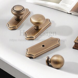 May include: Antique brass cabinet knobs and pulls. The knobs have a round shape, and the pulls have a rectangular backplate with decorative edges. The hardware is made of metal and has a vintage aesthetic.