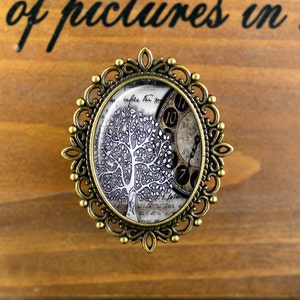 Puede incluir: Un broche ovalado de bronce con un diseño ornamentado. El broche presenta un camafeo de vidrio con una imagen en blanco y negro de un árbol y una esfera de reloj.