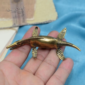 May include: A gold-colored metal figurine of a prehistoric marine reptile, a plesiosaur, with a long neck and flippers. The figurine is detailed with textured scales and is held in a hand.