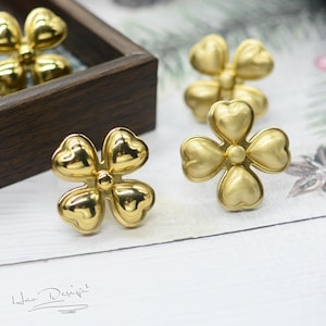 Puede incluir: Cuatro adornos de metal de color dorado en forma de flor. Las flores tienen cinco pétalos cada una y un centro elevado. Las flores están dispuestas sobre una superficie de madera clara.