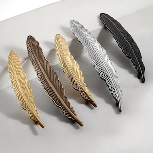 Op de afbeelding: Een verzameling decoratieve meubelgrepen in de vorm van veren, in diverse metallic afwerkingen. De grepen zijn goud, brons, zilver en zwart. Elke greep heeft een gedetailleerd verenontwerp, wat elegantie toevoegt.