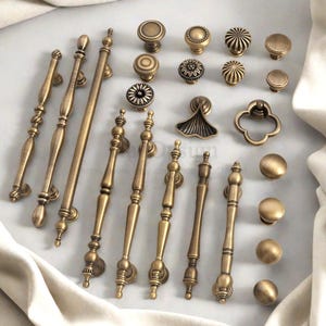 May include: A collection of antique brass cabinet hardware, including knobs and pulls. The hardware features various designs, such as floral patterns, rounded shapes, and elongated bar pulls. The hardware is arranged on a light-coloured surface.