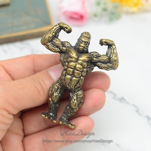 以下が含まれることがあります： 二頭筋を曲げているゴリラの金色の金属製フィギュア。ゴリラは平らな表面に立っており、詳細なテクスチャ加工が施されています。