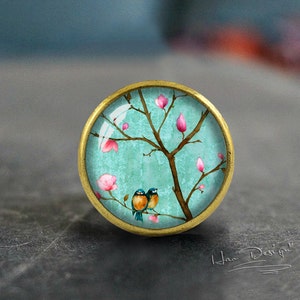 Könnte beinhalten: Ein runder, messingfarbener Knopf mit einem Glaseinsatz, der ein gemaltes Design eines Baumes mit rosa Blüten und zwei blauen Vögeln zeigt. Der Hintergrund ist hellblau.