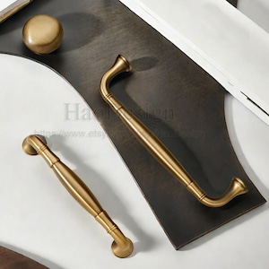 May include: A collection of antique brass cabinet hardware, including two pull handles and a round knob. The handles have a curved design and are displayed on a dark brown surface. The hardware has a vintage aesthetic.