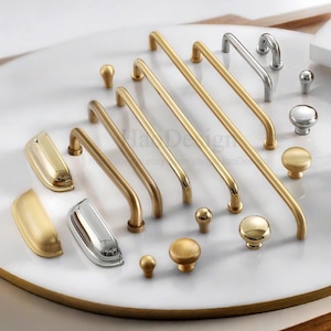 Pode incluir: Uma coleção de ferragens para armários em acabamentos dourados e prateados. A variedade inclui vários estilos de puxadores, incluindo puxadores de barra e puxadores de concha, juntamente com vários designs de botões. O hardware é exibido em uma superfície branca.