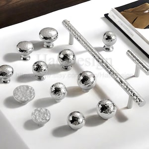 Puede incluir: Un conjunto de herrajes plateados para armarios, que incluyen pomos y tiradores con textura martillada. La imagen muestra varias formas y tamaños sobre una superficie blanca. Acabado metálico reflectante, ideal para muebles de cocina o baño.