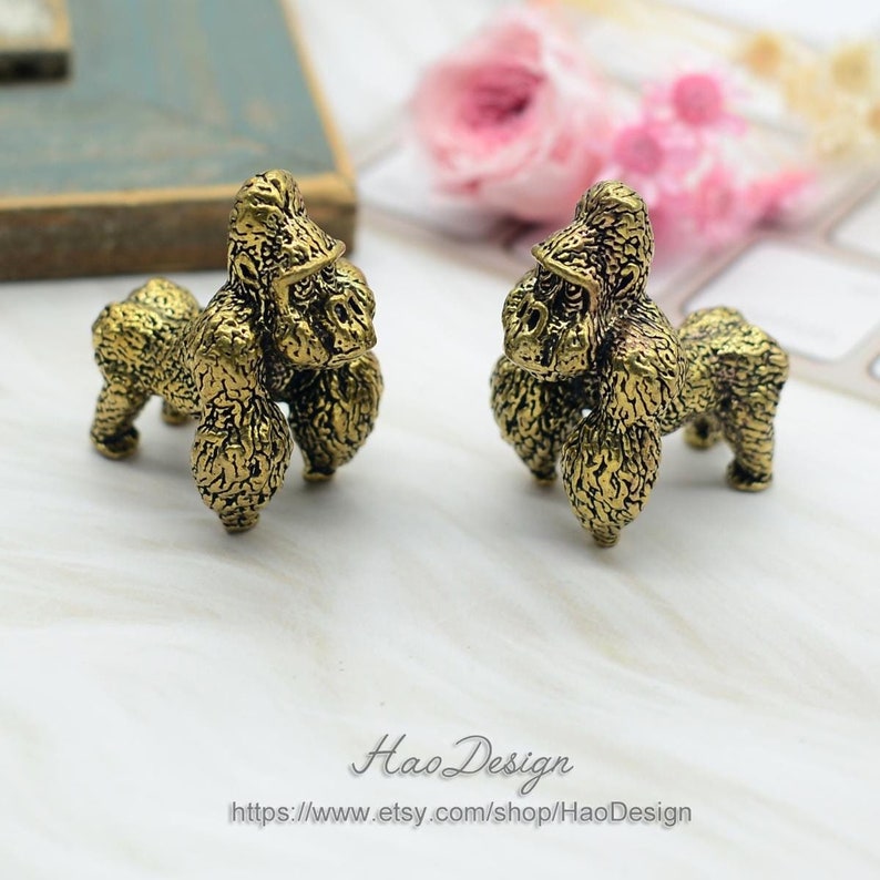 May include: Two small gold-colored gorilla figurines. The figurines are detailed and have a textured surface. They are standing on a white surface.