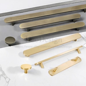 May include: A selection of brass-coloured cabinet handles and knobs. The image shows long bar pulls, shorter pulls, and round knobs with a brushed finish. The hardware is displayed on a light surface. The Etsy shop name and URL are visible.
