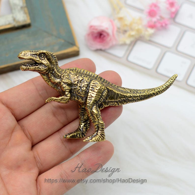 Pode incluir: Uma figurinha de dinossauro Tyrannosaurus Rex de metal dourado. A figurinha &eacute; detalhada com escamas texturizadas e tem uma cauda longa.