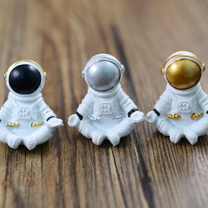 May include: Three white astronaut figurines in a seated yoga pose. Each astronaut has a different colored helmet: black, silver, and gold. The figurines have gold accents on the hands and feet.