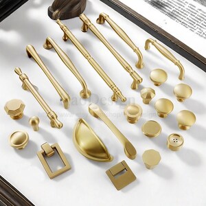 May include: A collection of gold-toned cabinet hardware, including various handles, knobs, and pulls. The hardware features different shapes and designs, such as bar pulls, cup pulls, and octagonal knobs. The items are arranged on a white surface.