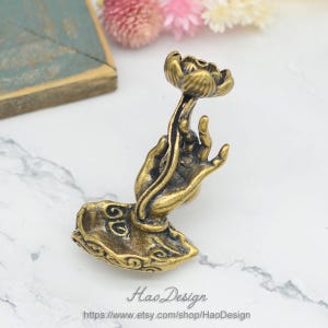 Puede incluir: Un quemador de incienso de metal dorado en forma de mano sosteniendo una flor de loto. La mano descansa sobre una base en forma de nube.