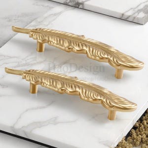 Puede incluir: Dos tiradores dorados en forma de pluma. Los tiradores son de metal y tienen un diseño detallado de pluma. Los tiradores se muestran sobre una superficie de mármol blanco. El texto "HaoDesign" es visible.