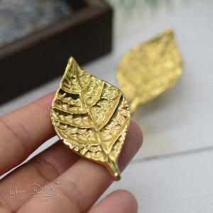 May include: A pair of gold leaf-shaped earrings. The leaves have a textured surface and are detailed with veins.