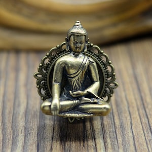 May include: A gold-toned metal ring featuring a small, detailed sculpture of a seated Buddha. The Buddha is in a meditative pose with hands resting in the lap. The ring is set in a decorative, ornate frame.