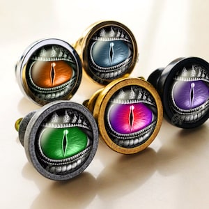 Może przedstawiać: Pięć okrągłych gałek do szafek w srebrnym, złotym i czarnym wykończeniu. Każda gałka przedstawia szczegółową, czarno-białą ilustrację oka smoka z innym kolorem tęczówki: niebieskim, pomarańczowym, fioletowym, różowym i zielonym.