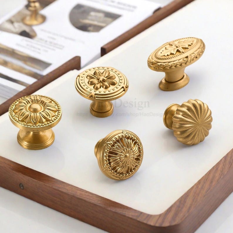 K&ouml;nnte beinhalten: Eine Sammlung von f&uuml;nf goldfarbenen Schrankkn&ouml;pfen mit verschiedenen dekorativen Designs. Die Kn&ouml;pfe weisen florale und geometrische Muster mit runden und ovalen Formen auf. Sie werden auf einer wei&szlig;en Oberfl&auml;che mit einem Holzrahmen pr&auml;sentiert.