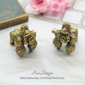 May include: Two gold-toned metal cabinet knobs shaped like poodles. The knobs have a detailed, textured finish.