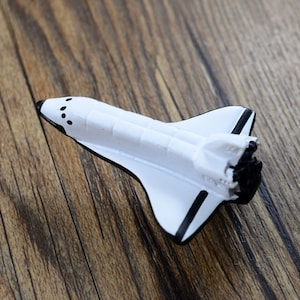 May include: White toy space shuttle with black details. The shuttle has a pointed nose with three small black circles and a black stripe on the side.