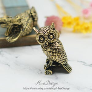 Puede incluir: Una figurita de búho de color dorado con plumas detalladas y ojos grandes. El búho está posado en una rama y tiene una superficie texturizada.