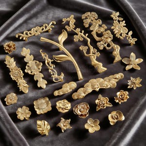 May include: A collection of gold-colored cabinet hardware, including handles and knobs. The designs feature floral, leaf, and branch motifs. The hardware is arranged on a dark gray velvet surface.