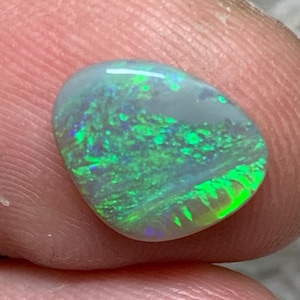 May include: A polished opal gemstone with a rounded shape. The opal displays a vibrant play of colour, with flashes of green and blue against a translucent background. The gemstone is held between fingers.
