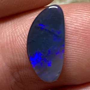May include: A polished, oval-shaped opal gemstone. The opal has a dark blue base colour with vibrant flashes of blue and purple throughout. The stone is held between two fingers.