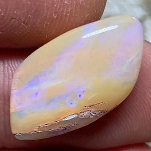 Op de afbeelding: Een marquisevormige cabochon opaal edelsteen met een glad, gepolijst oppervlak met een licht perzik- en crèmekleurige basis. De steen vertoont iriserende flitsen van lichtpaars en blauw kleurenspel over het gehele oppervlak.