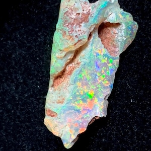 Könnte beinhalten: Ein roher, unpolierter Opal-Edelstein mit einer porösen, rotbraunen Matrix-Basis, der lebhafte, mehrfarbige Lichtreflexe in Grün, Gelb, Orange und Blau über seine unregelmäßige, texturierte Oberfläche und gezackten Kanten verteilt.