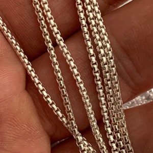 Op de afbeelding: Een close-up van een zilveren kettingketting. De ketting is gemaakt van kleine, vierkante schakels en is erg delicaat.