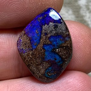 Könnte beinhalten: Ein polierter, tropfenförmiger Boulder-Opal-Edelstein mit leuchtenden, irisierenden blauen und violetten Blitzen vor einer dunklen, texturierten braunen Eisensteinmatrix. Die Oberfläche des Steins ist glatt und reflektierend und zeigt ein natürliches Farbspiel.