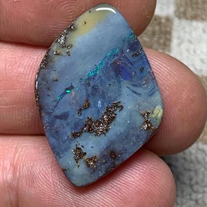 Könnte beinhalten: Ein polierter, unregelmäßig geformter Opal-Edelstein. Der Stein weist eine Mischung aus blauen, grünen und gelben Farbtönen mit dunkelbraunen Einschlüssen auf. Der Opal wird zwischen den Fingern gehalten und zeigt seine natürliche Schönheit und sein einzigartiges Muster.
