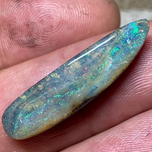 Könnte beinhalten: Ein polierter Opal-Edelstein mit länglicher Form. Der Opal zeigt ein Farbspiel, darunter Grün, Blau und rote Reflexe, wodurch ein schillernder Glanz entsteht. Der Stein wird in einer Hand gehalten, was seine natürliche Schönheit hervorhebt.