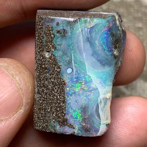May include: A rectangular boulder opal gemstone featuring a dark brown ironstone matrix on the left side and a vibrant, iridescent play-of-color on the right. The surface displays swirling patterns of electric blue, green, and purple flashes.