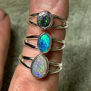 Op de afbeelding: Drie zilveren ringen met ovale opalen. De bovenste ring heeft een donkergrijze opaal, de middelste ring heeft een blauwgroene opaal en de onderste ring heeft een witte opaal met regenboogkleuren.