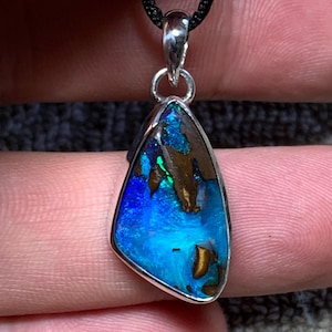 Pode incluir: Um pingente com uma opala boulder de formato irregular em gota, com reflexos iridescentes azuis e verdes vibrantes, engastada numa moldura de prata polida. A pedra apresenta inclusões de matriz castanha natural e está suspensa num cordão preto.