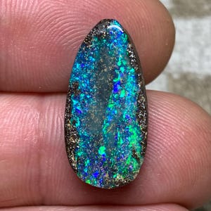 Op de afbeelding: Een traanvormige boulder-opaal edelsteen met een donkerbruine matrixbasis en levendige, iriserende flitsen van elektrisch blauw en heldergroen kleurenspel over het gepolijste oppervlak, met een natuurlijke, onregelmatige textuur.