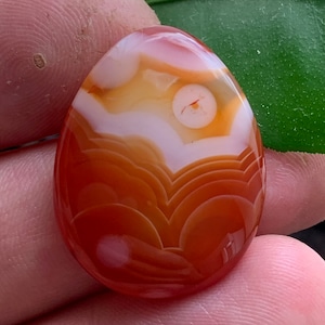 Könnte beinhalten: Ein glatter, ovaler Karneol-Stein mit einem weißen und orangefarbenen Achatmuster. Der Stein hat eine rotbraune Farbe mit weißen Bändern und einer weißen Mitte.