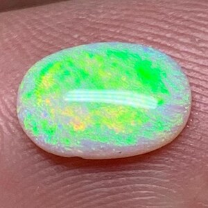 Peut inclure: Une pierre d'opale ovale aux couleurs vives, affichant des nuances de vert, de jaune et de blanc. La surface polie de l'opale révèle un jeu de couleurs irisé. La pierre est tenue entre les doigts, mettant en valeur sa beauté naturelle.