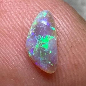 Peut inclure: Une petite pierre précieuse opale en forme de goutte avec une base translucide, présentant des éclats vibrants de vert néon, de bleu électrique et de magenta qui jouent sur sa surface polie, révélant un motif irisé distinct.