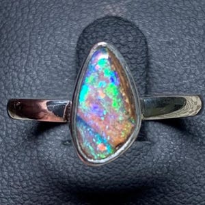 Op de afbeelding: Een zilveren ring met een druppelvormige opaal die een kleurenspel vertoont met levendige flitsen van groen, blauw en oranje. De gepolijste metalen band houdt de steen vast in een eenvoudige zetting, wat de natuurlijke textuur benadrukt.