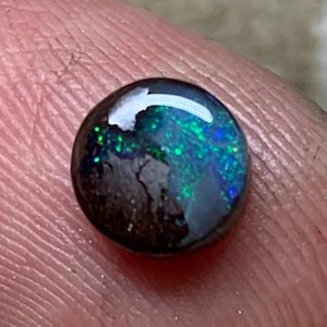 Peut inclure: Une petite pierre précieuse opale noire, ronde et polie, présentant une couleur de base sombre avec des éclats iridescents vibrants de lumière verte et bleue sur sa surface. La pierre a une forme cabochon lisse et bombée avec une finition brillante.