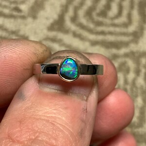 May include: A silver ring with a pear-shaped opal gemstone. The opal displays a vibrant play of color, with flashes of blue and green. The ring has a simple, modern design, showcasing the gemstone's natural beauty.